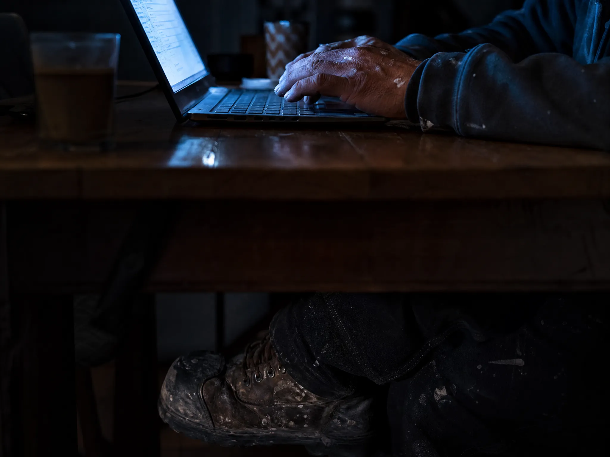 Trades worker at the kitchen table late at night, lit by laptop glow, still doing paperwork
